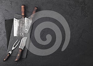 Vintage meat knife and fork and hatchets with stone chopping board and black table background. Butcher utensils. Space for text