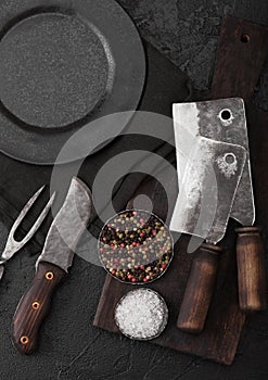 Vintage meat knife and fork and hatchets with vintage chopping board and plate on black table background. Butcher utensils. Close