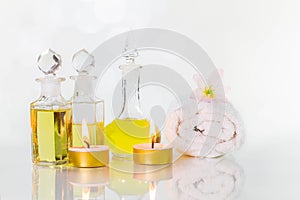 Vintage old bottles of aromatic oils with burned candles, flowers and white towel on glossy white table on white background