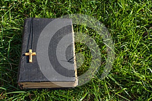 Vintage old holy bible book, grunge textured cover with wooden christian cross. Retro styled image on grass background.