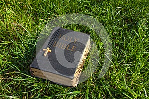 Vintage old holy bible book, grunge textured cover with wooden christian cross. Retro styled image on grass background.