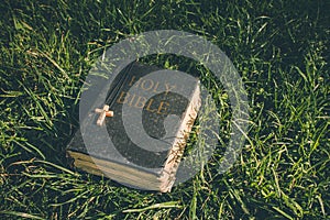 Vintage old holy bible book, grunge textured cover with wooden christian cross. Retro styled image on grass background.