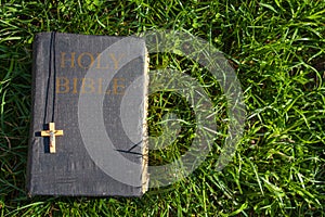 Vintage old holy bible book, grunge textured cover with wooden christian cross. Retro styled image on grass background.