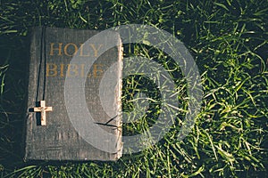 Vintage old holy bible book, grunge textured cover with wooden christian cross. Retro styled image on grass background.