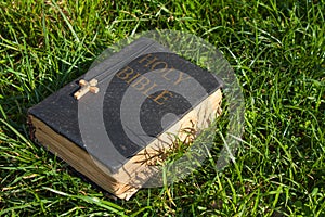 Vintage old holy bible book, grunge textured cover with wooden christian cross. Retro styled image on grass background.