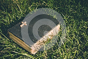 Vintage old holy bible book, grunge textured cover with wooden christian cross. Retro styled image on grass background.