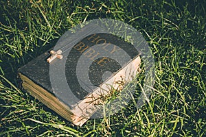 Vintage old holy bible book, grunge textured cover with wooden christian cross. Retro styled image on grass background.