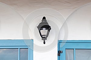 Vintage old street classic black iron outdoor lantern, lamp on white house wall between two blue window frames