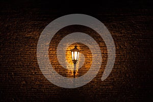 Vintage old street lantern lit with an orange light in front of a brick wall during a gloomy dark night..