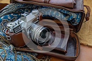 A vintage photo camera and a brown leather bag with scarf on sack cloth background