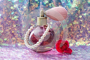 Vintage pink color glass perfume bottle with pearls and red rose blossom on shiny purple sequin texture.