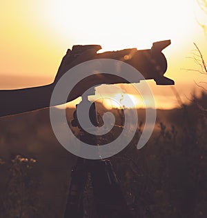 Vintage silhouette of a professional camera on a tripod in a field at sunset with lens hood