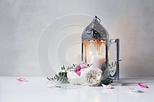 Vintage silver Moroccan, Arabic lantern with glowing candle, green branches, rose flowers and pink petals on white table
