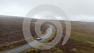 Vintage sports car driving on wet road winding between grasslands in countryside. Cloudy day in hazy landscape. Ireland