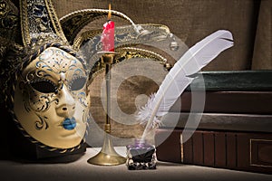 Vintage still life with old books near inkstand, feather, venezian mask and burning red candle in candlestick on canvas background