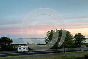 A vintage style camper van, motorhome with a metal trunk attached to its roof parked on a seafront road at sunset