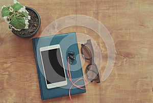 Vintage style, white smart phone with black blank space on the screen, glasses. earphones and notebook on wooden background.