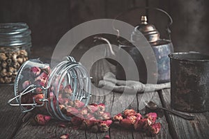 Vintage teapot, cup of healthy herbal tea, and glass jars of dry rose buds and daisy tea.