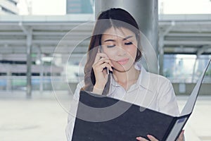 Vintage toned image of attractive young Asian secretary talking on phone and holding document folder on walkway in city