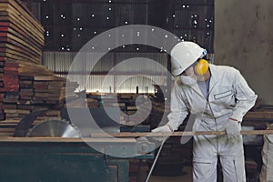 Vintage toned image of professional young Asian worker in white uniform and safety equipment cutting a piece of wood on table saw