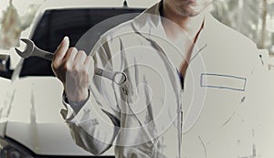 Vintage toned image of professional young mechanic man in uniform holding wrench against car at the repair garage.