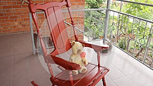 A vintage toy bear sits and sways in a rocking chair on an open veranda. Summer greens.