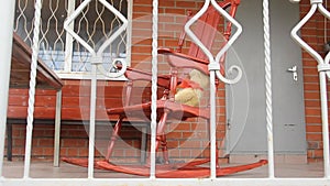 A vintage toy bear sits and sways in a rocking chair on an open veranda. Summer greens. Shooting through the railing