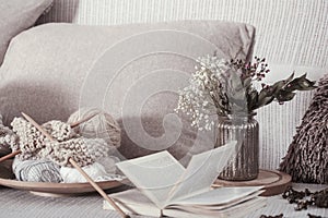 Vintage wooden knitting needles and threads on a cozy sofa with pillows and a vase of flowers. Open book for reading