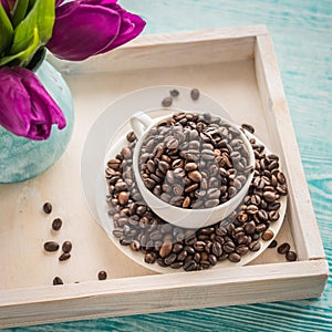 Vintage wooden tray with porcelain cup full of coffee beans and pink flowers on shabby chic mint background, top view