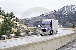 Violet bonnet big rig semi truck with dry van semi trailer driving with cargo on winding road with mountain pass in California