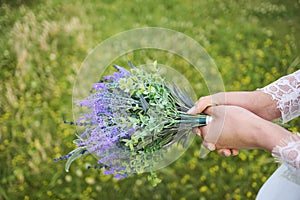 Violet bouquet held tightly in hands of a woman in white dress on field of yellow blossoms.