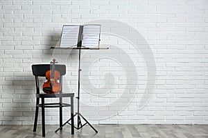 Violin, chair and note stand with music sheets near brick wall.