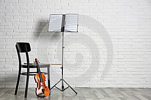 Violin, chair and note stand with music sheets near brick wall.