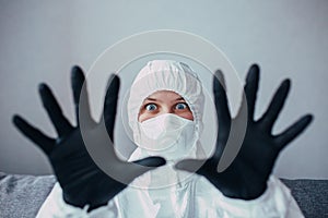 A virologist in a white protective suit with a black mask and medical gloves with fear on face shows hands into the camera. Doctor