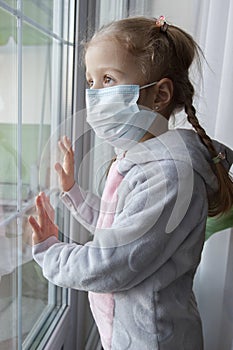 Virus concept, little girl in face mask looking through window at home or clinic. Portrait of sad kid during quarantine due to