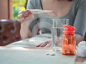 Virus protection concept, glass of water, orange bottle of pills in blurry background of a woman looking at thermometer, protect