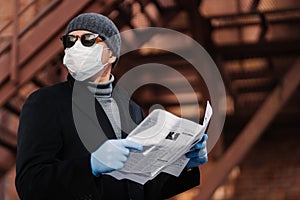 Virus spread and quarantine concept. Serious man wears sunglasses, hygienic mask and rubber gloves, looks away, reads press,