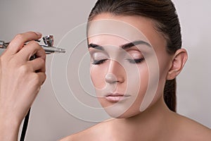 Visagist making makeup for model with aerograph, isolated on grey. Background with hand holding aerograph. Visagist using aerograp photo