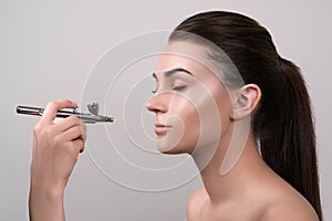 Visagist making makeup for model with aerograph, isolated on grey. Background with hand holding aerograph. Visagist using aerograp photo