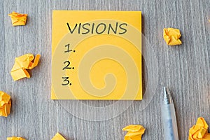 Visions word on yellow note with Businessman holding pen and crumbled paper on wooden table background. Business Goals, Mission,