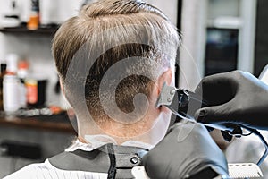 Visit to the barbershop. Hairdresser, barber shaves client`s temples,sides with hair clipper. Young stylish man makes fashionable