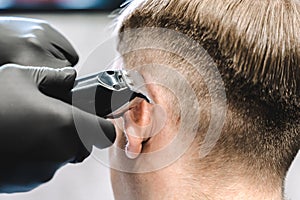 Visit to the barbershop. Hairdresser, barber shaves client`s temples,sides with hair clipper. Young stylish man makes fashionable
