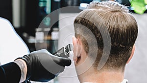 Visit to the barbershop. Hairdresser, barber shaves client`s temples,sides with hair clipper. Young stylish man makes fashionable
