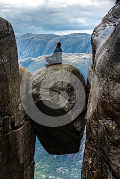 Visiting Norway Kjeragbolten located south of Lysefjorden Young woman sits on top of gian boulder in meditative pose