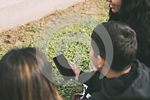 vista desde atras grupo de latinos riendo sentados en el suelo en un parque con un smartphone photo