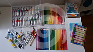 Art supplies, including paint tubes and a rainbow of colored pencils, are neatly displayed on a white table.