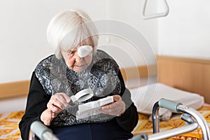 Visually impaired elderly 95 years old woman sitting at the bad trying to read with magnifying glass.