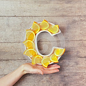 Vitamin C nutrient in food concept. Woman hand holding plate in shape of letter C with orange slices on wooden background. Flat