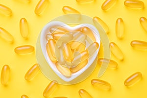 Vitamin D and Omega 3 fish oil capsules supplement in a heart-shaped plate on yellow background. Concept of healthcare