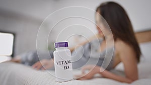 Vitamin D3 bottle on bed with blurred pregnant woman reading book lying at background. Brunette Caucasian young